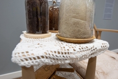 Están Aquí Installation Detail, 2024-26, repurposed object Gossip chair, specialty fabric, Botonaduras, artist’s grandmother’s wool cards, brown felt, antique white fringe trim, clear glass cloche domes, gray, red & brunette hair, fabric arches, hand-sewn human hair, patch on unprimed canvas, 34 3/4” x 36” x 17”, installed at Martin Museum of Art, Waco, Texas, 2026